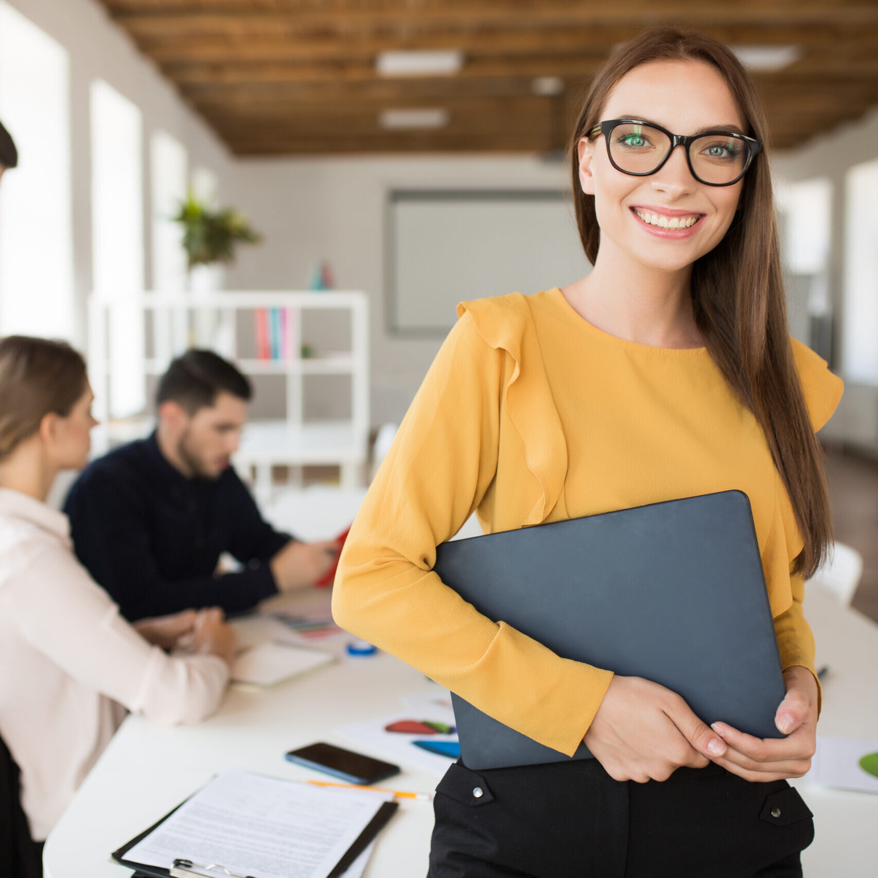Young smiling business woman in eyeglasses happily looking in camera holding folder in hands in office with colleagues on background